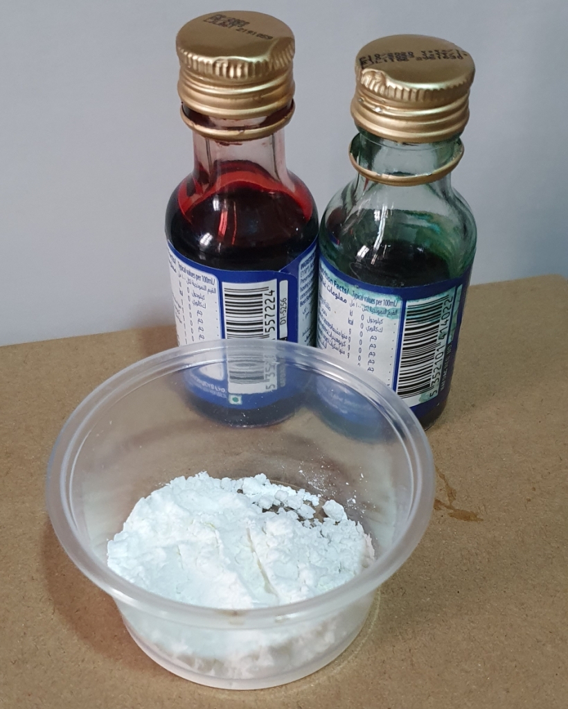 Image of a bottles of red and blue food colouring and a table spoon of cornstarch in a transparent container.